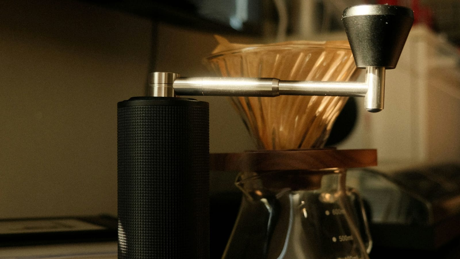 Moedor manual de café moderno, de metal, ao lado de um dripper sobre uma jarra de vidro