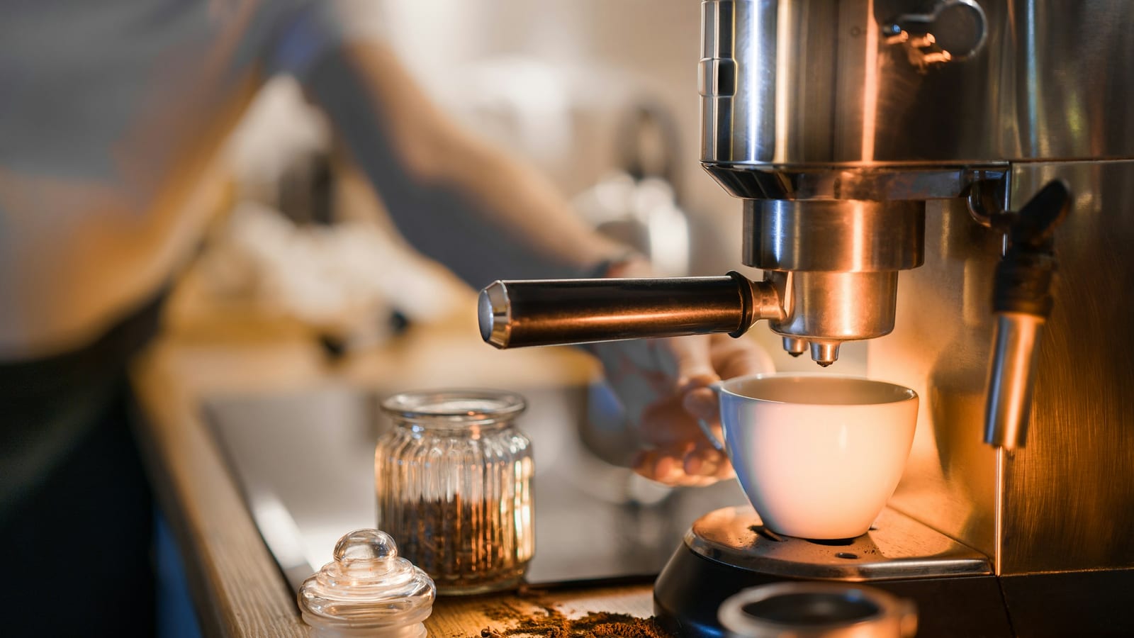 Máquina de espresso com porta-filtro enchendo uma xícara branca, em uma cozinha com luz quente