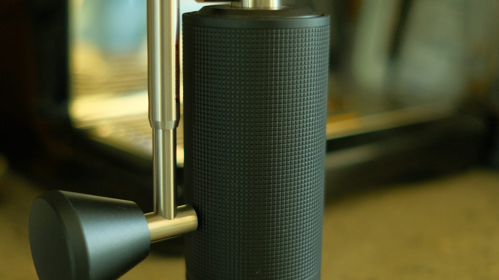 Moedor manual de café de corpo texturizado e manivela de metal, em frente a uma máquina de espresso