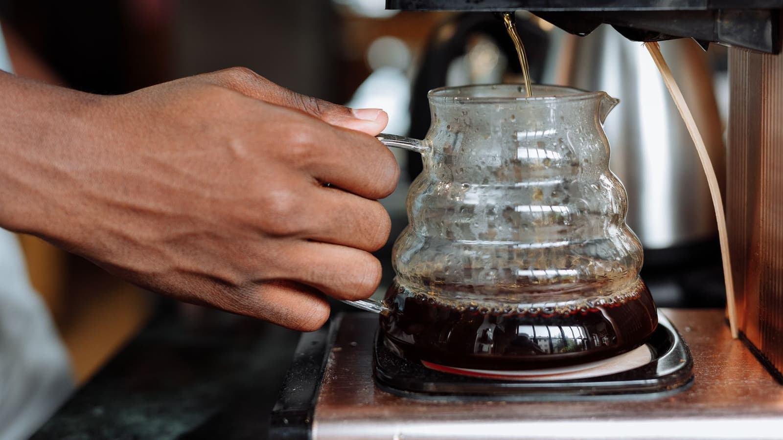 Cafeteira elétrica de filtro pingando café coado em uma jarra de vidro, com a mão segurando a alça