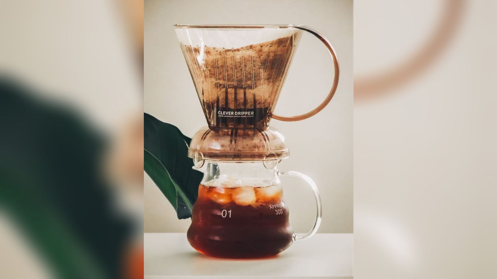 Clever Dripper apoiado sobre uma jarra de vidro com café, em fundo claro com uma folha ao lado