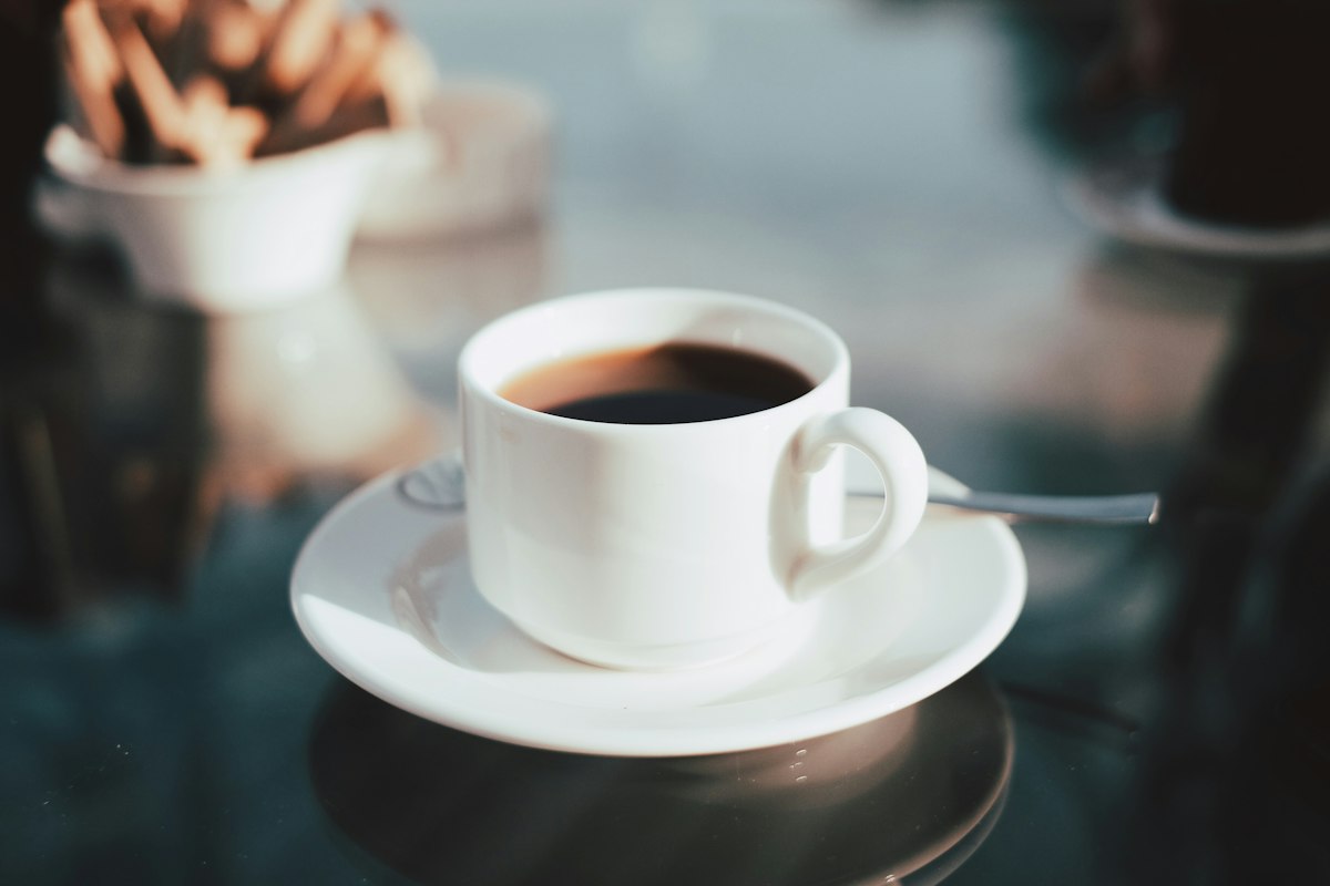 Xícara branca com café preto sobre pires, em uma mesa