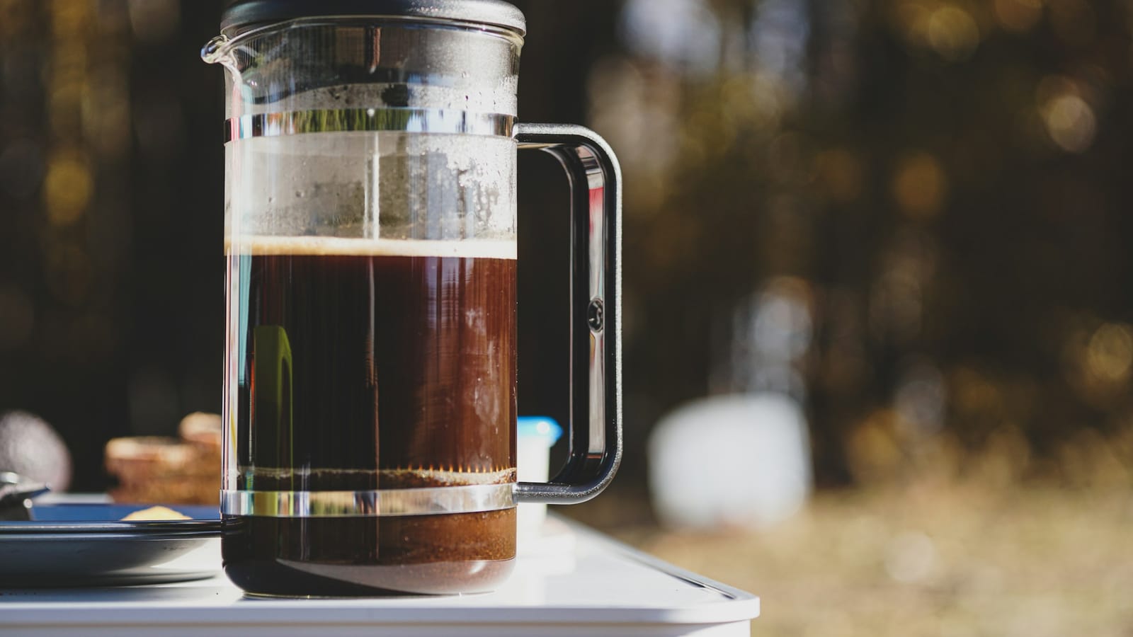 Prensa francesa de vidro com café pronto e o êmbolo levantado, sobre uma mesa ao ar livre