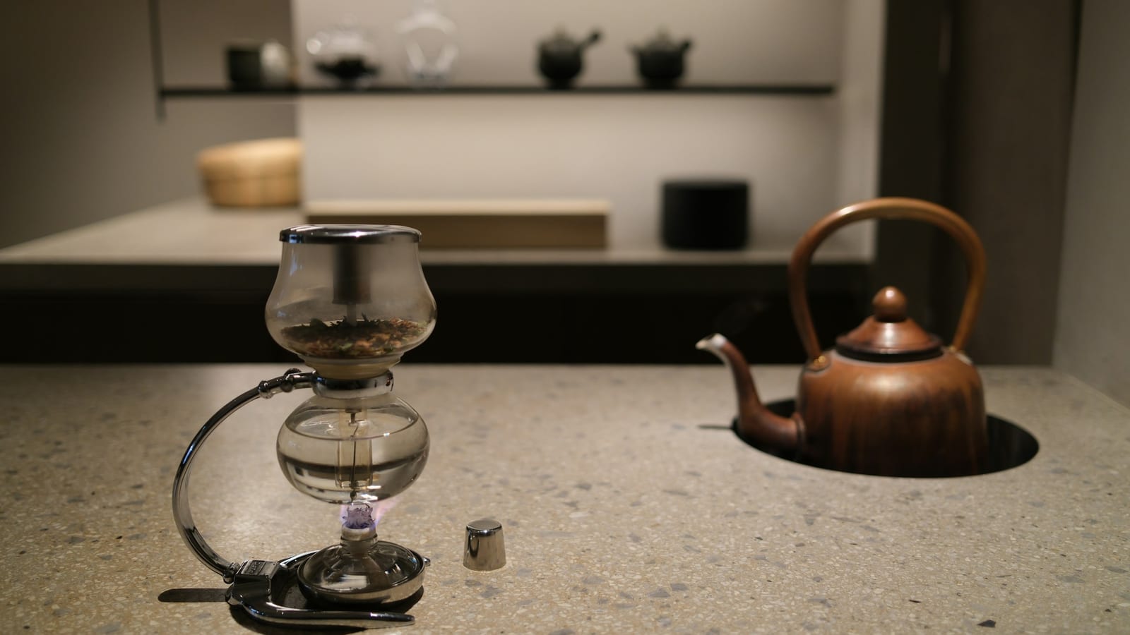 Sifão de vidro para café sobre uma bancada de pedra, com uma chaleira de cobre ao fundo