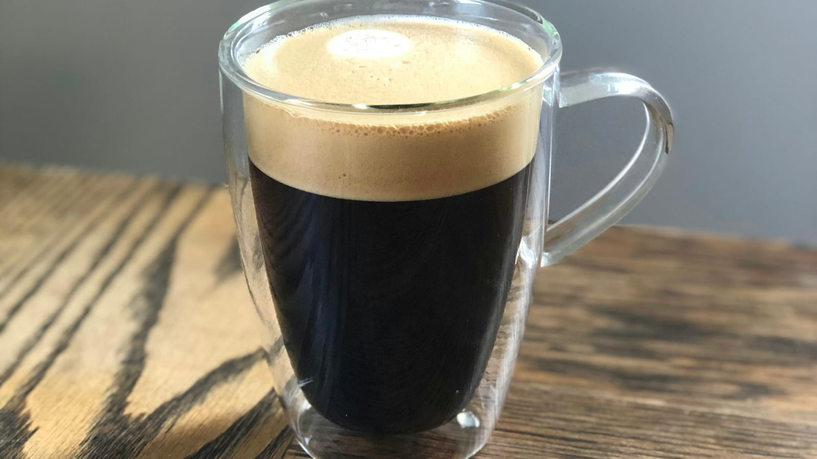 Caneca de vidro de parede dupla com café preto e crema clara, sobre mesa de madeira