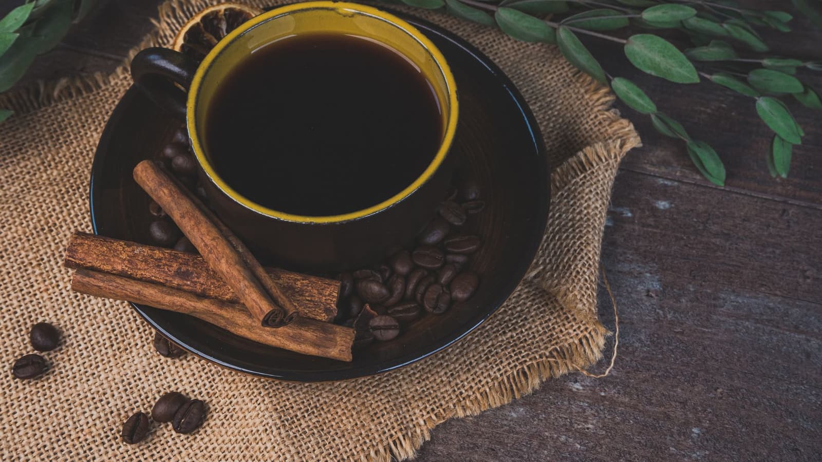 Xícara de café preto sobre um pires escuro com paus de canela, grãos de café e um pano de juta, em mesa rústica de madeira