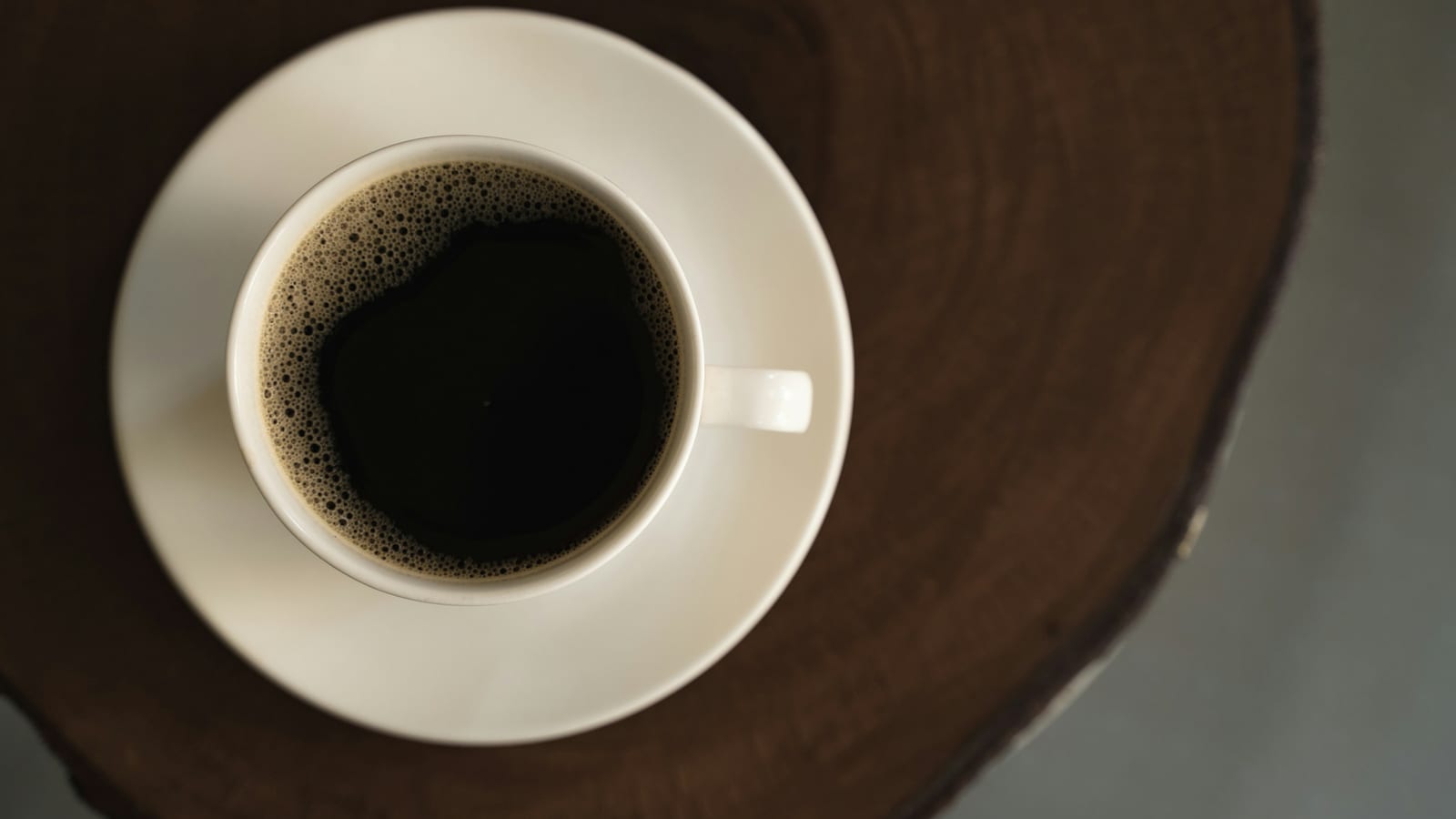 Xícara branca com café preto coado sobre pires e uma base de madeira, vista de cima
