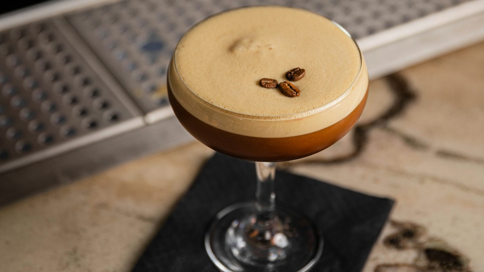 Espresso martini em taça coupe com espuma cremosa e três grãos de café, sobre um balcão de bar