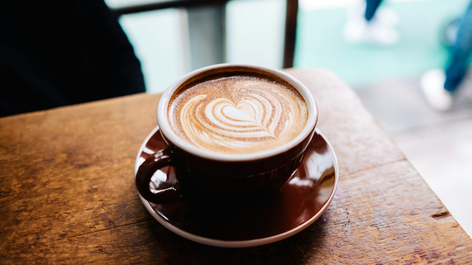 Xícara marrom sobre pires com latte e arte de coração na espuma, sobre uma mesa de madeira