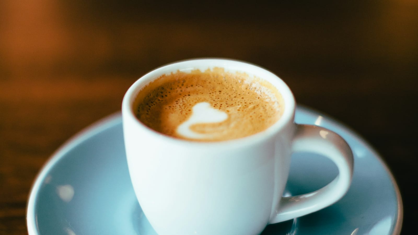 Xícara pequena branca com espresso e um coração de espuma de leite, sobre pires azul