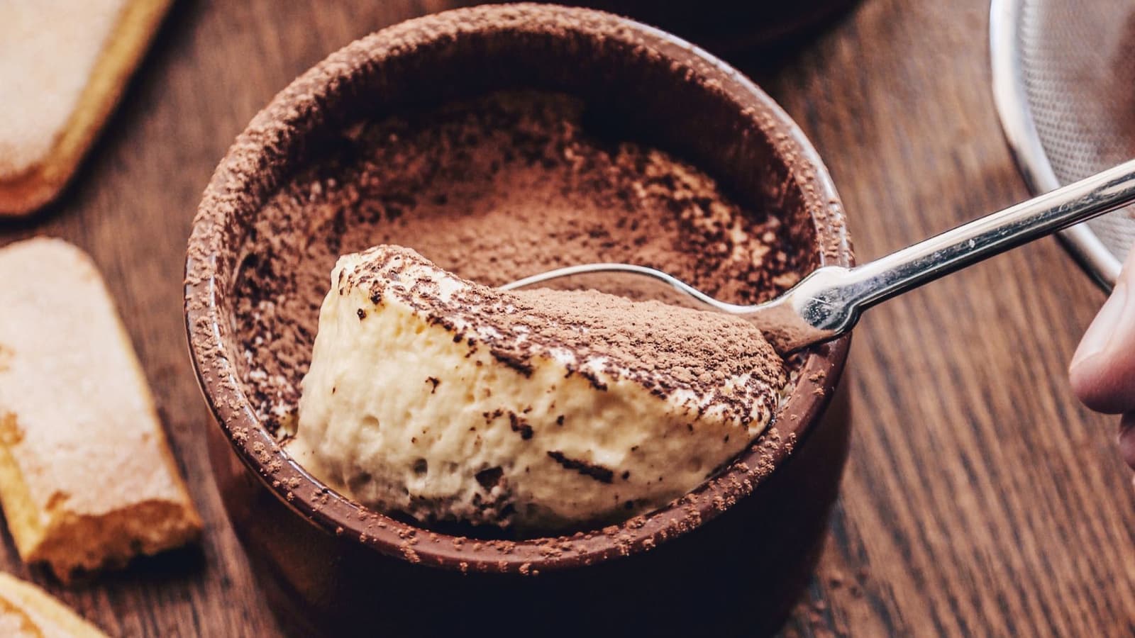 Colherada de mousse cremosa e aerada em um ramequim de barro polvilhado com cacau, sobre mesa de madeira com biscoitos champanhe